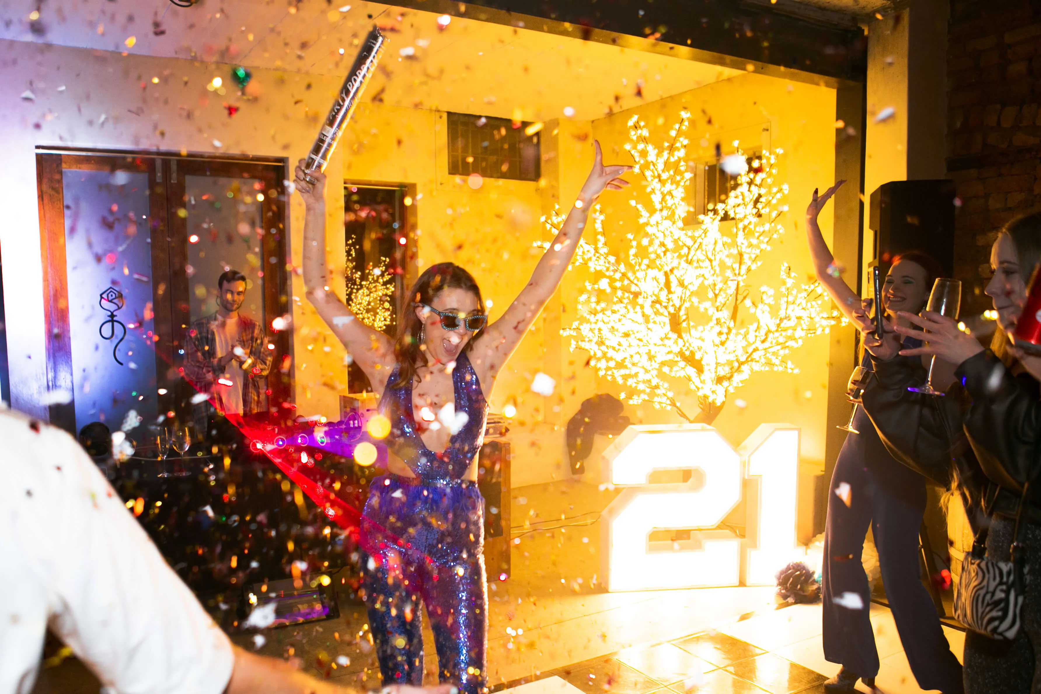 Woman in a sequinned jumpsuit raising a confetti cannon beside light-up 21 letters at a 21st birthday party