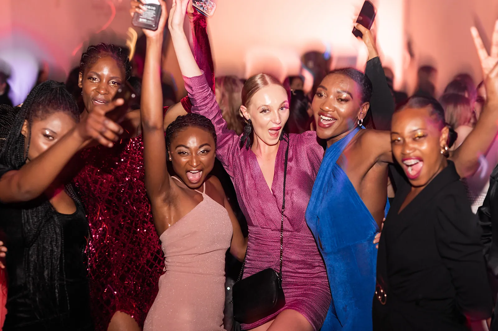 Six women in party dresses cheering with their arms raised and phones in hand on a dance floor