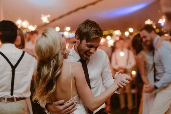 Groom smiling as he dances with his bride under string lights in a marquee