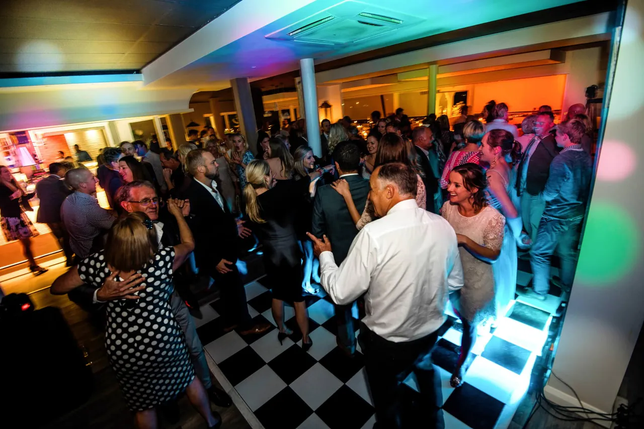 A packed dance floor under coloured lights at a wedding, guests dancing on black and white tiles