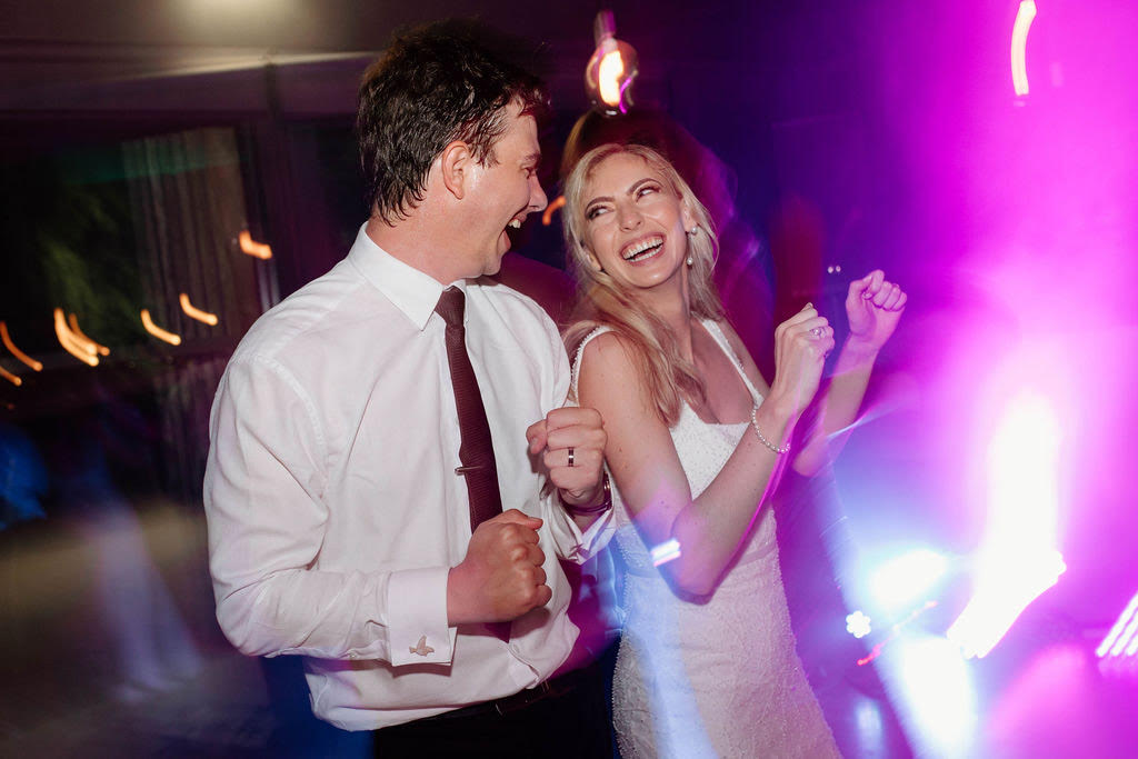Bride and groom laughing as they dance together under pink and violet party lights